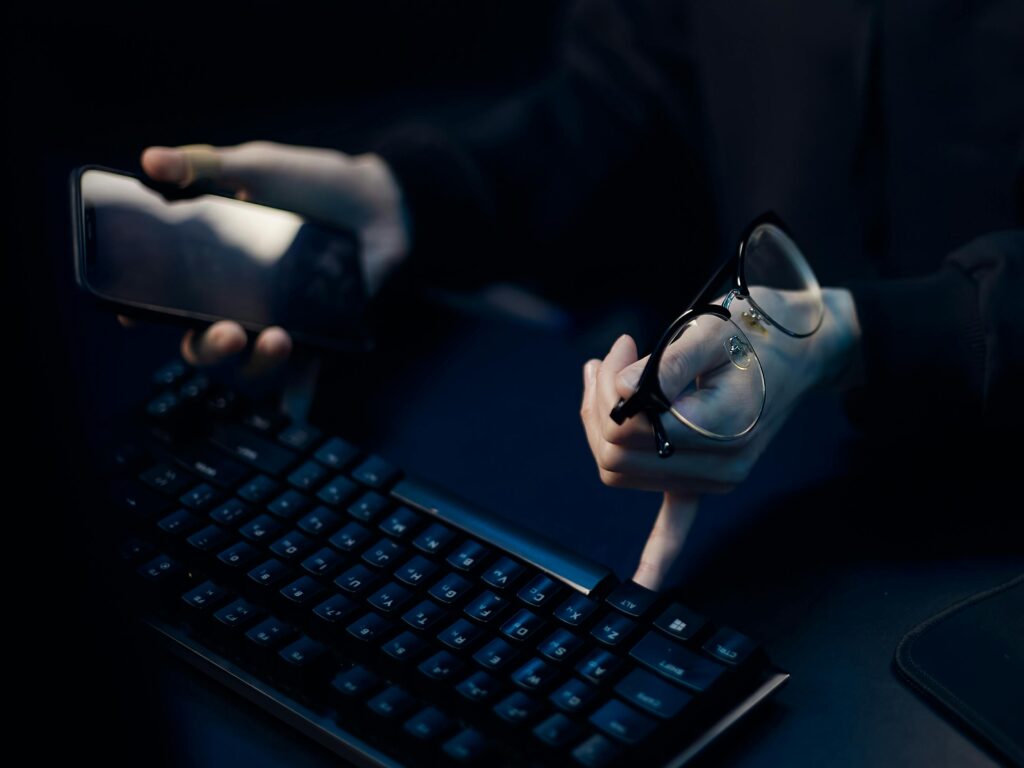Close-up of hands using smartphone and computer keyboard, holding eyeglasses, focused workspace.